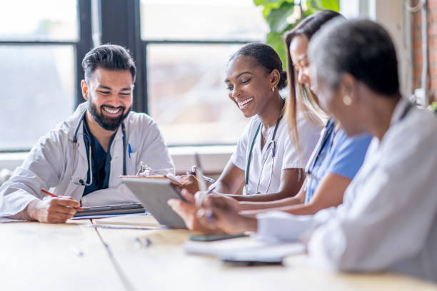 A small group of four medical professionals sit around a boardroom table as they meet to discuss patient cases.  They are each dressed professionally in scrubs and lab coats as they focus on working together.